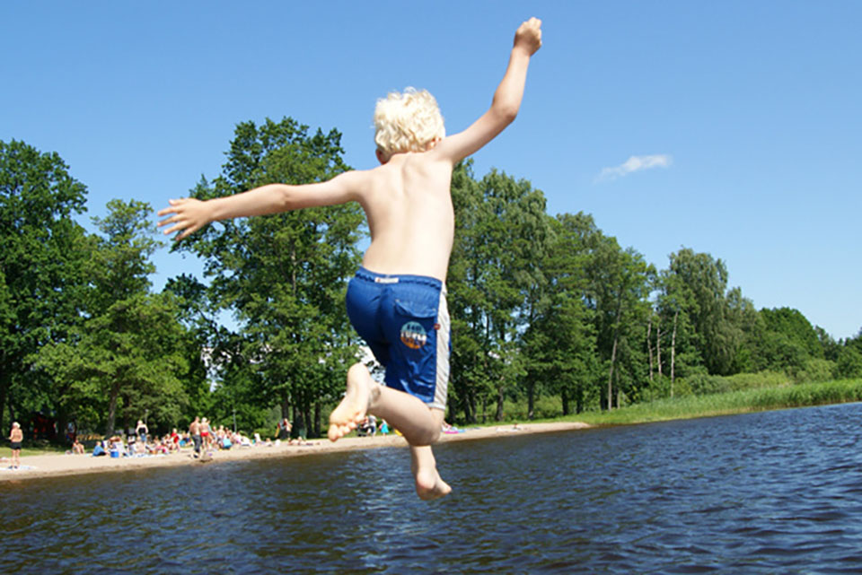 en ung pojke som hoppar i vattnet. I bakgrunden ser man människor på en strand.
