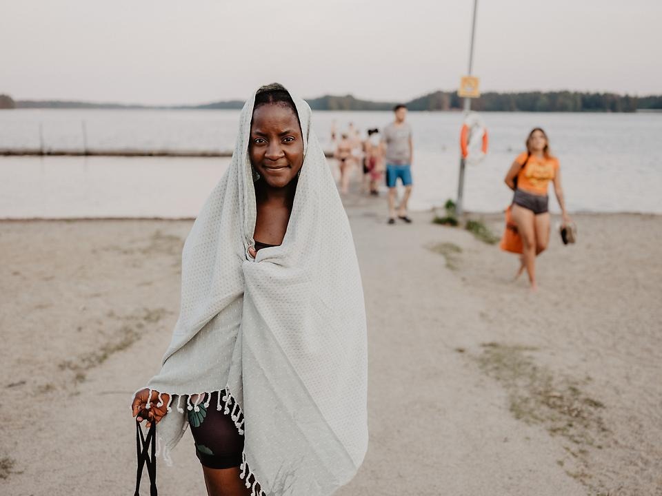 Ung kvinna i handduk och badkläder står framför en badbrygga med människor vid en sjö.