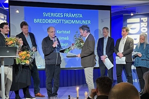 En scen med fem män och en kvinna. I mitten håller en man i ett diplom, en bukett blommor och en mikrofon. På en skärm bakom står det Sveriges främsta breddidrottskommuner: Karlstad, Umeå, Växjö.